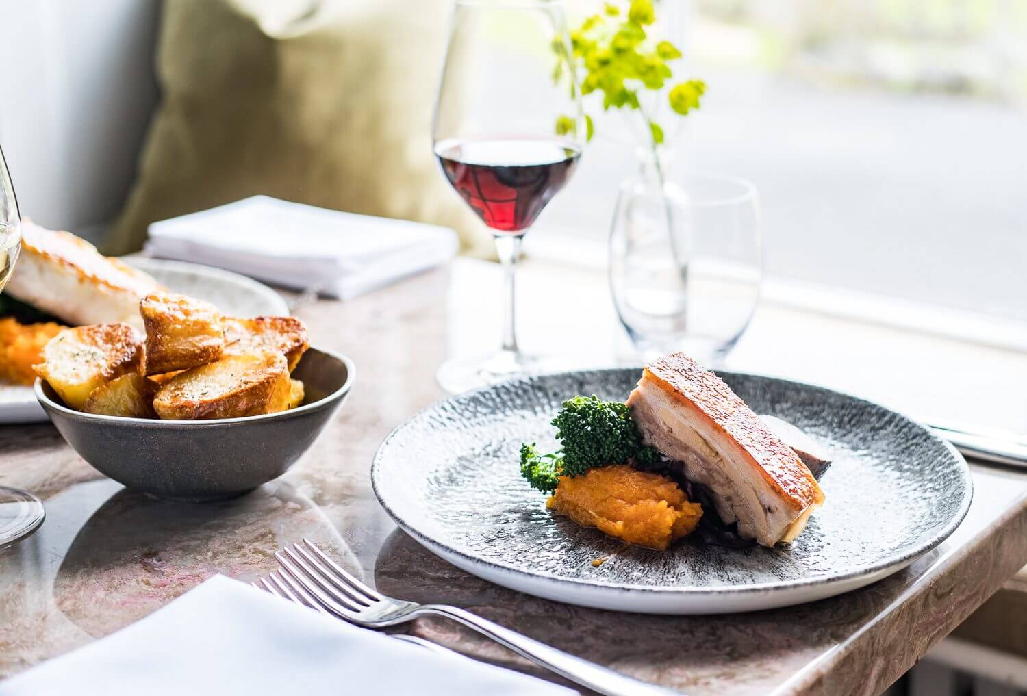 Belly pork roast on a bed of seasonal vegetables with a side of roast potatoes in a bowl